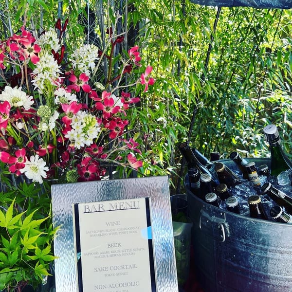 Outdoor bar setup with a "BAR MENU" sign, beer bottles in an ice bucket, and floral arrangements.