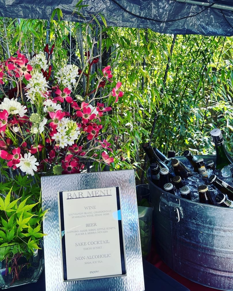 Outdoor bar setup with a "BAR MENU" sign, beer bottles in an ice bucket, and floral arrangements.