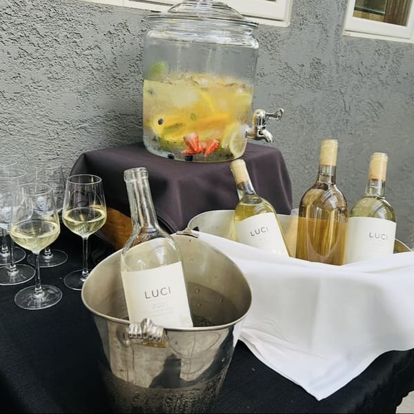 A beverage station with a fruit-infused drink dispenser, wine bottles in ice buckets, and wine glasses.