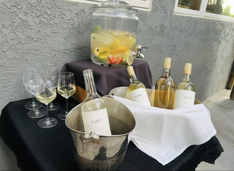 A beverage station with a fruit-infused drink dispenser, wine bottles in ice buckets, and wine glasses.