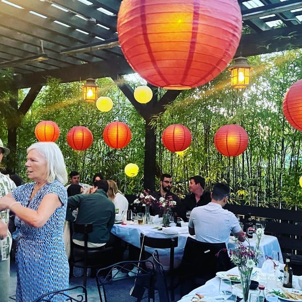 People dining outdoors under a pergola adorned with red and yellow paper lanterns, surrounded by bamboo.
