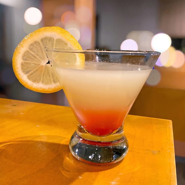 A vibrant cocktail with a red bottom and white top, garnished with a lemon slice, on a yellow surface.