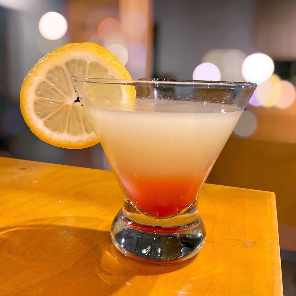 A vibrant cocktail with a red bottom and white top, garnished with a lemon slice, on a yellow surface.