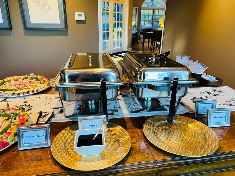 Catering buffet with assorted sushi platters and chafing dishes for tempura, rice, and chicken teriyaki