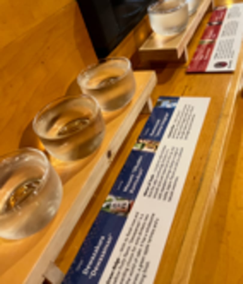 Sake tasting flight with three glasses and descriptive cards on a wooden bar.
