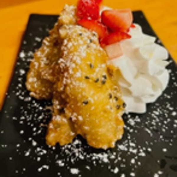 Fried banana dessert with powdered sugar, sesame seeds, strawberries, and whipped cream on a black plate.