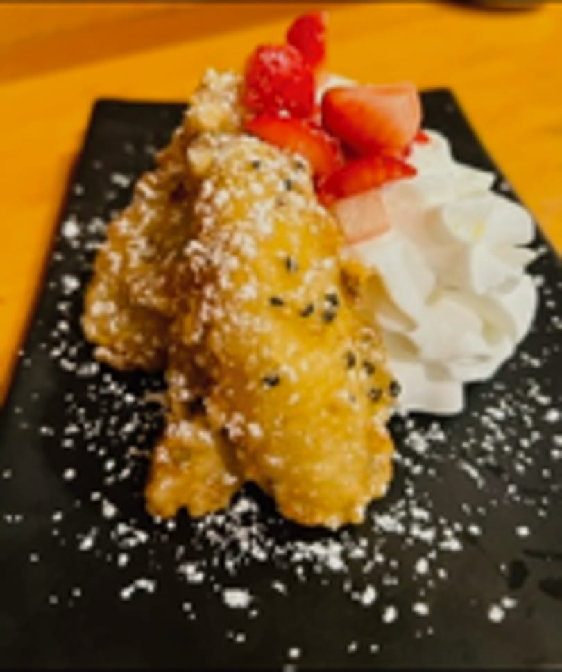 Golden fried bananas, powdered sugar, black sesame, strawberries, and whipped cream on a black plate.