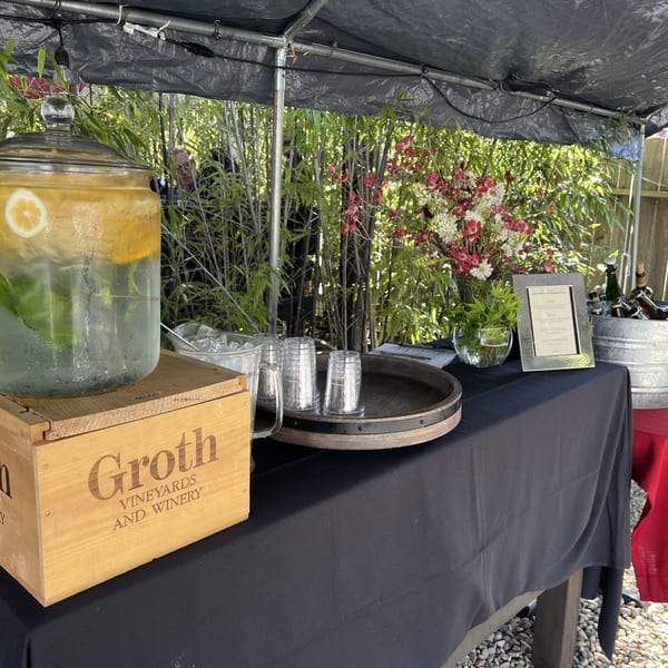 Outdoor beverage station with lemon water, beer in a bucket, and flowers under a black canopy.