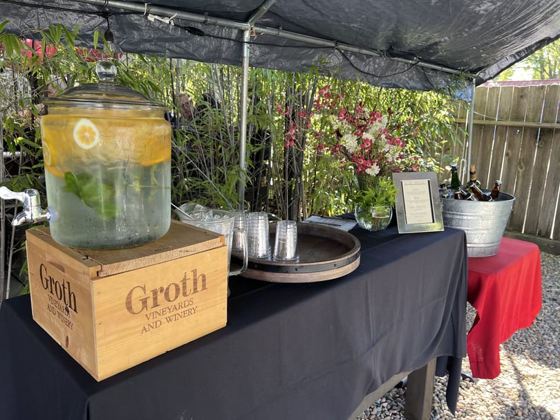 Outdoor beverage station with lemon water, beer in a bucket, and flowers under a black canopy.