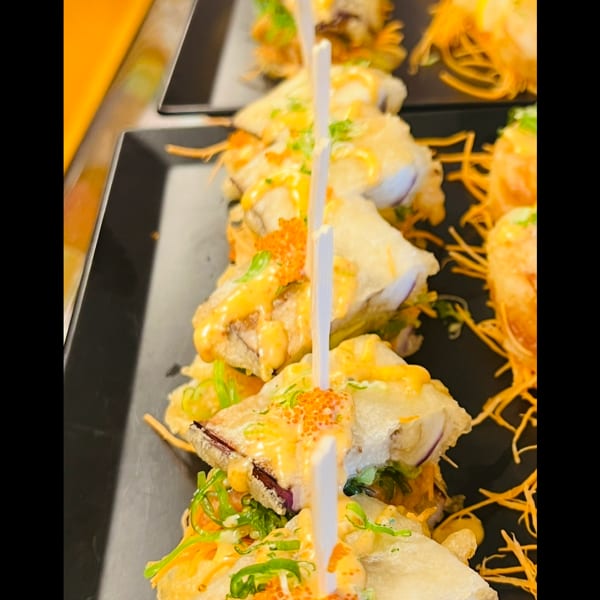 Tempura-fried sushi pieces with sauce, roe, and green onions, held by skewers on a black tray.