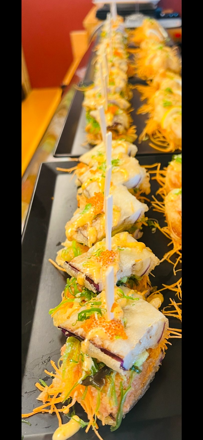 Tempura-fried sushi pieces with sauce, roe, and green onions, held by skewers on a black tray.