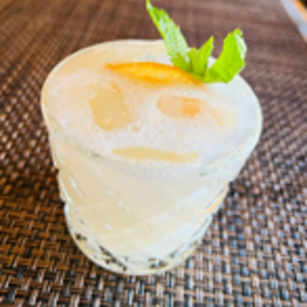 A foamy cocktail garnished with a mint sprig and orange peel on a woven placemat.