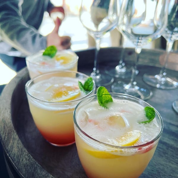 Three vibrant cocktails with lemon slices and mint on a wooden tray, with wine glasses in the background.