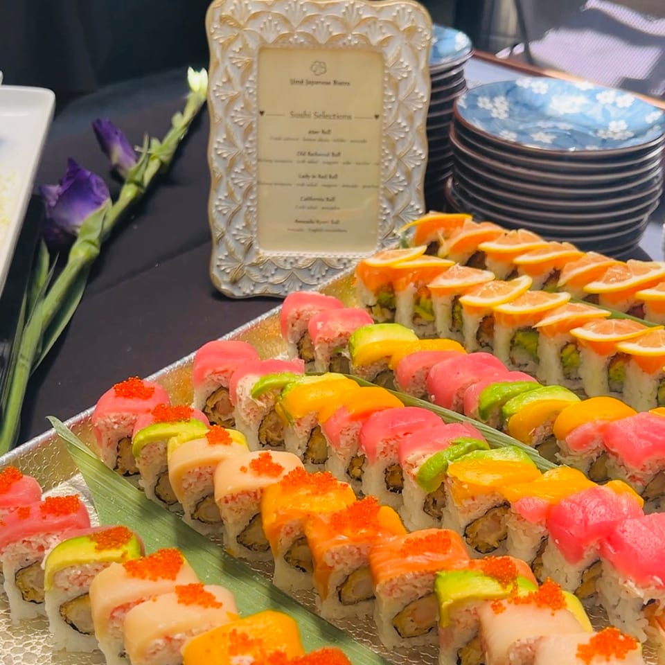 Colorful sushi rolls with various toppings on a buffet table with a menu and stacked plates.