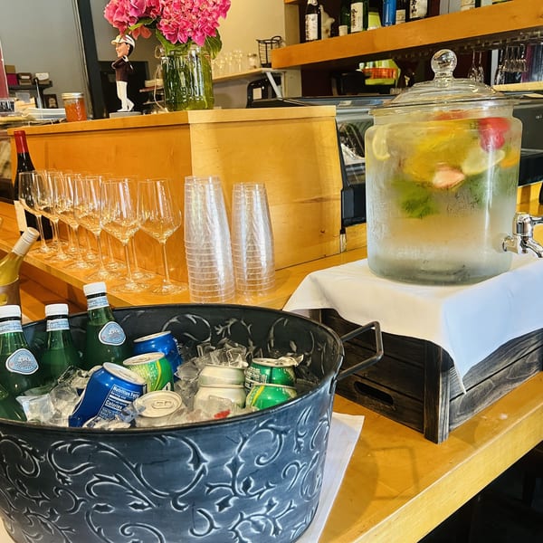 A beverage station with a drink dispenser, wine glasses, and a bucket of bottled drinks on a wooden counter.