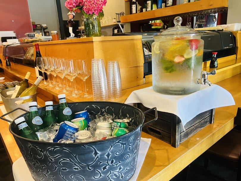 A beverage station with a drink dispenser, wine glasses, and a bucket of bottled drinks on a wooden counter.