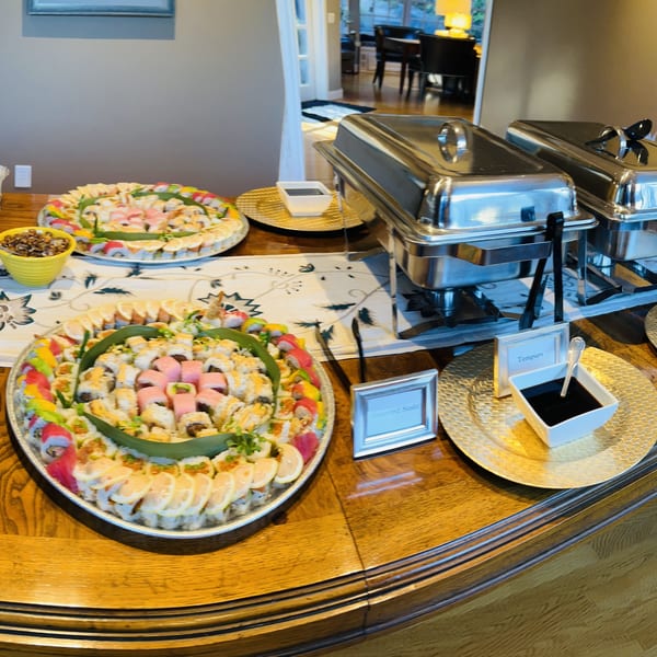 A buffet table laden with sushi platters, egg rolls, salad, and chafing dishes.