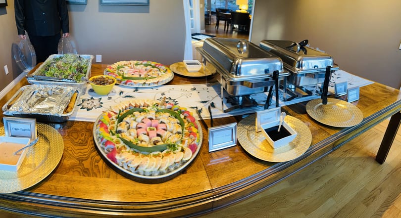A buffet table laden with sushi platters, egg rolls, salad, and chafing dishes.