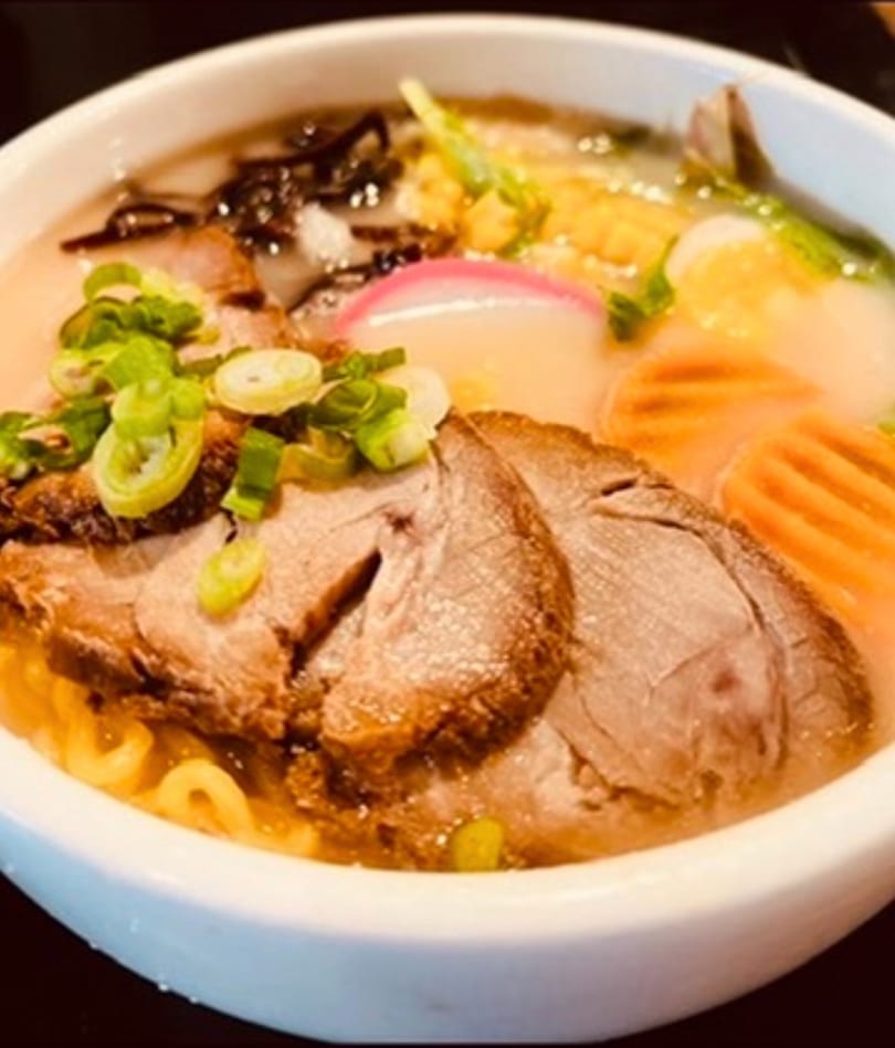 Close-up of a ramen bowl with sliced pork, noodles, corn, fish cake, and green onions.