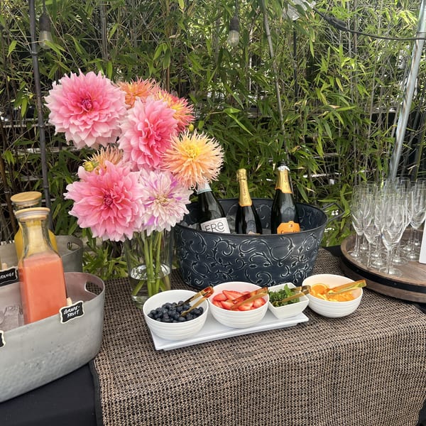 A Mom-osa bar with juices, champagne, fruit garnishes, and pink flowers against a bamboo backdrop.
