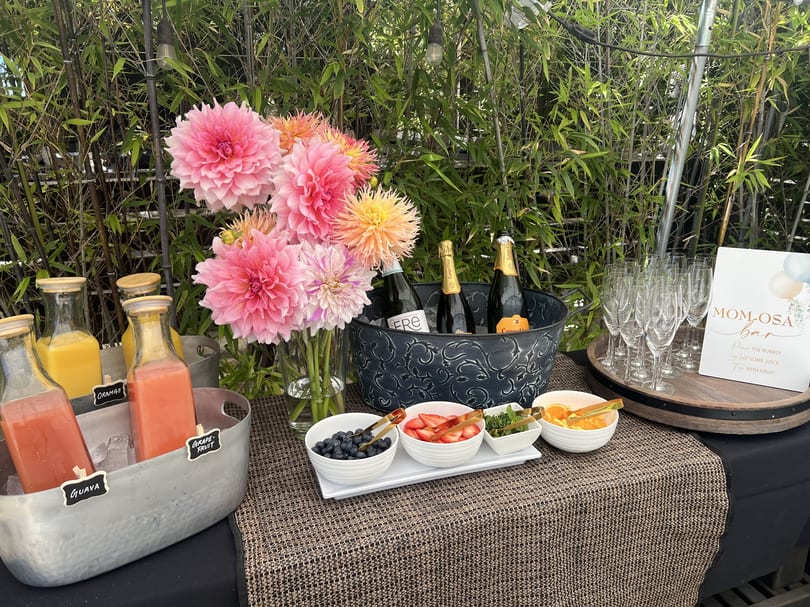 A Mom-osa bar with juices, champagne, fruit garnishes, and pink flowers against a bamboo backdrop.