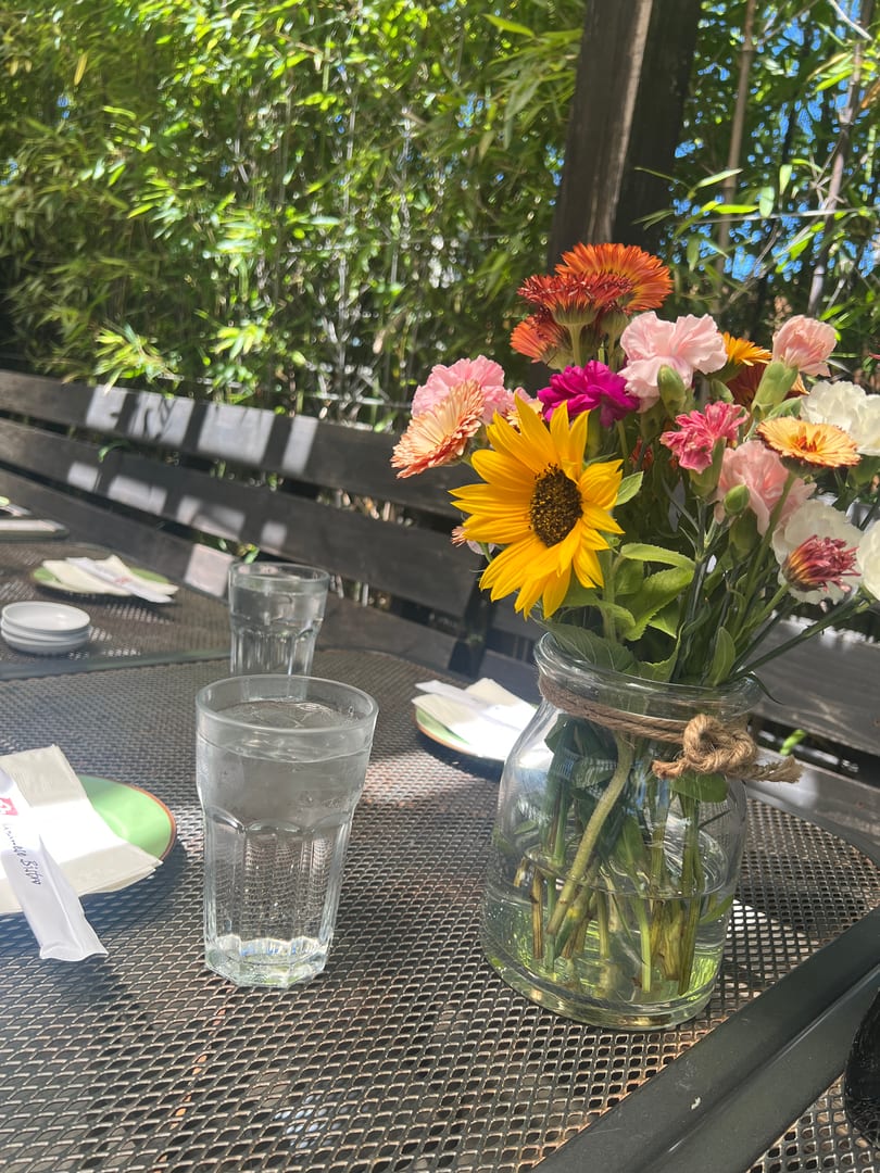 Outdoor dining table with a vibrant flower bouquet, water glasses, and plates under green foliage.